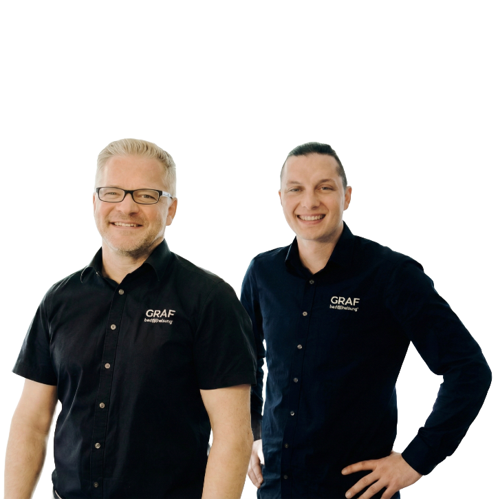 Zwei lächelnde Männer, die für Graf Patrick Manuel stehen, posieren vor einem schlichten weißen Hintergrund. Der Mann auf der linken Seite trägt ein kurzärmliges schwarzes Hemd und eine Brille. Der Mann auf der rechten Seite trägt ein langärmliges dunkelblaues Hemd. Beide Hemden zeigen das Logo "GRAF bad&heizung" auf der Brust.