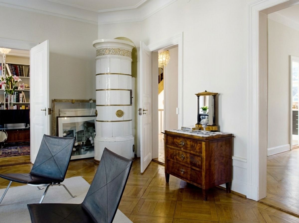Heller Raum mit Fischgrätparkett und einem hohen, zylindrischen, weißen Kachelofen mit goldenen Ringen und Medaillons. Zwei dunkelgraue Ledersessel stehen auf einem hellen Teppich. Links ist ein Wohnzimmer mit Bücherregalen sichtbar, rechts eine dunkle Holzkommode mit Spiegel und dahinter ein Flur mit Treppe.