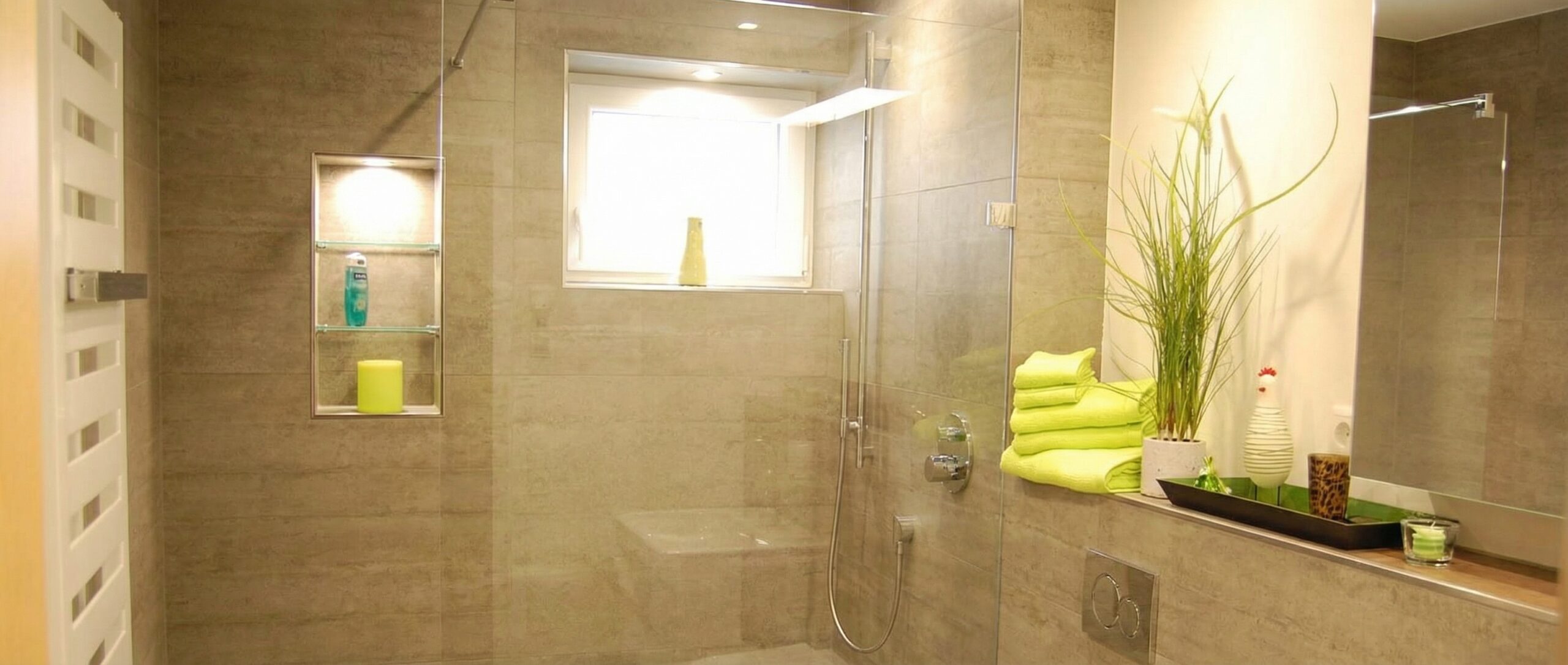 Modernes Badezimmer mit begehbarer Dusche, beige gefliesten Wänden und einem weißen Handtuchheizkörper. Die Glasduschkabine verfügt über eine Kopf- und Handbrause sowie eine beleuchtete Wandnische mit zwei Glasregalen, die Duschgel und eine gelbe Kerze halten. Über einer Ablagefläche in der Dusche ist ein Fenster mit einer gelben Vase. Rechts auf einer Ablage liegen hellgrüne Handtücher, eine grüne Topfpflanze und eine schwarze Dekoschale mit einer weißen Keramikhenne und einem Teelicht.