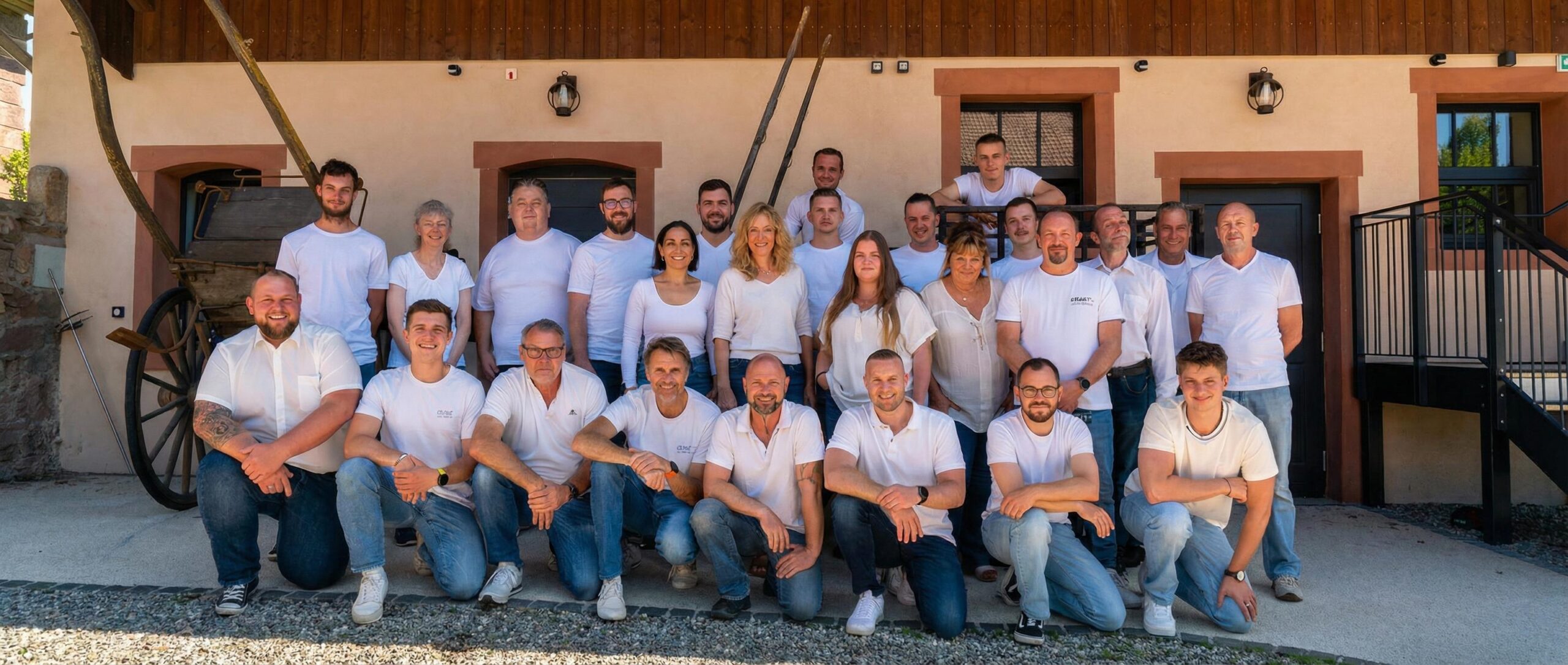 Eine große Gruppe von 25 Personen, bestehend aus Männern und Frauen unterschiedlichen Alters, posiert im Freien. Sie tragen alle weiße Oberteile und blaue Jeans. Die Gruppe ist in mehreren Reihen angeordnet, wobei einige Personen in der vorderen Reihe knien. Im Hintergrund ist ein hellbraunes Gebäude mit dunklen Holzverkleidungen im oberen Bereich sowie braunen Fenster- und Türrahmen sichtbar. Links neben der Gruppe steht ein alter, großer Holzwagen mit Speichenrädern und daneben lehnen landwirtschaftliche Werkzeuge an einer Steinmauer.